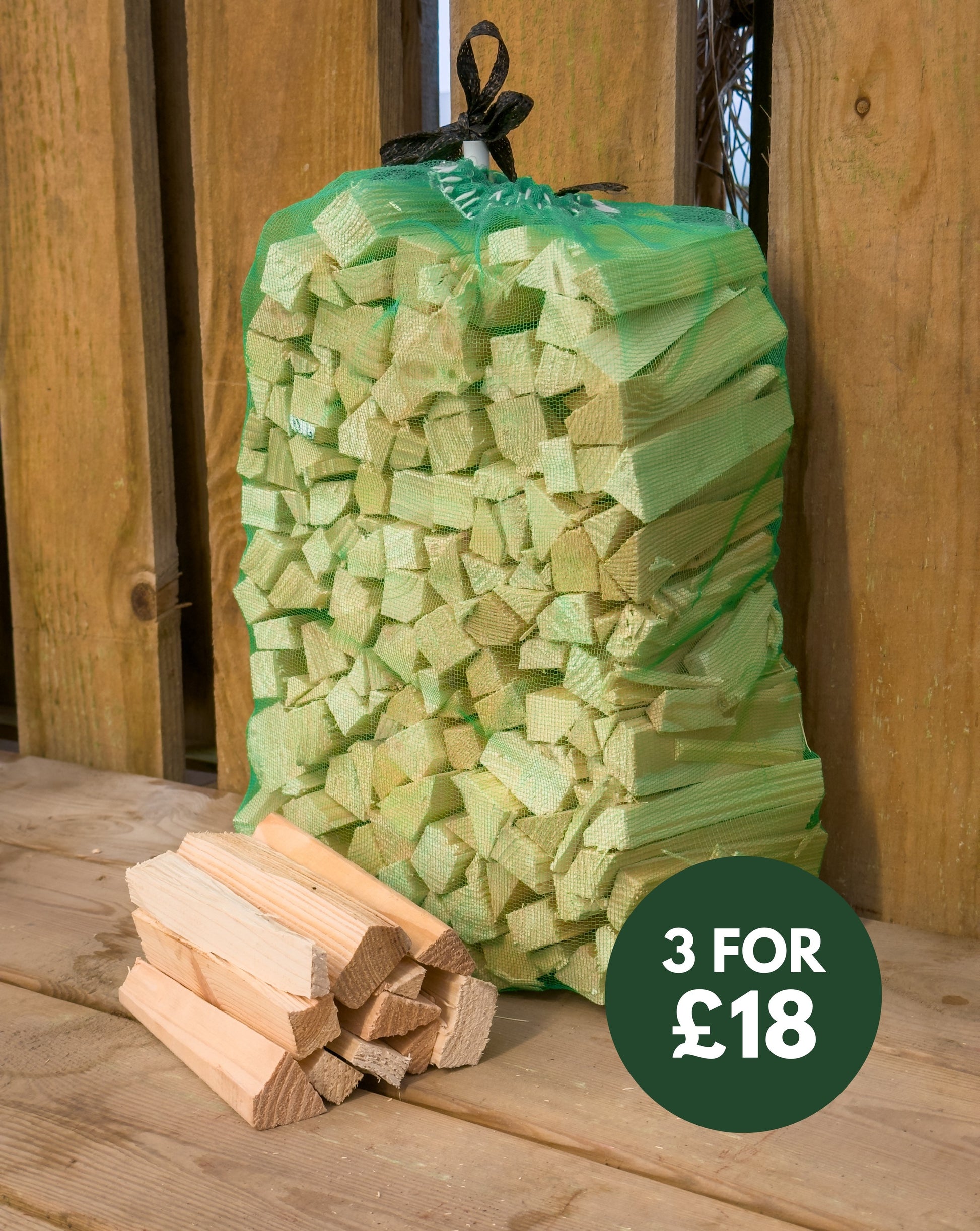 Net bag of kindling propped against fence, with loose split kindling sticks beside it and a badge reading 3 for £18.
