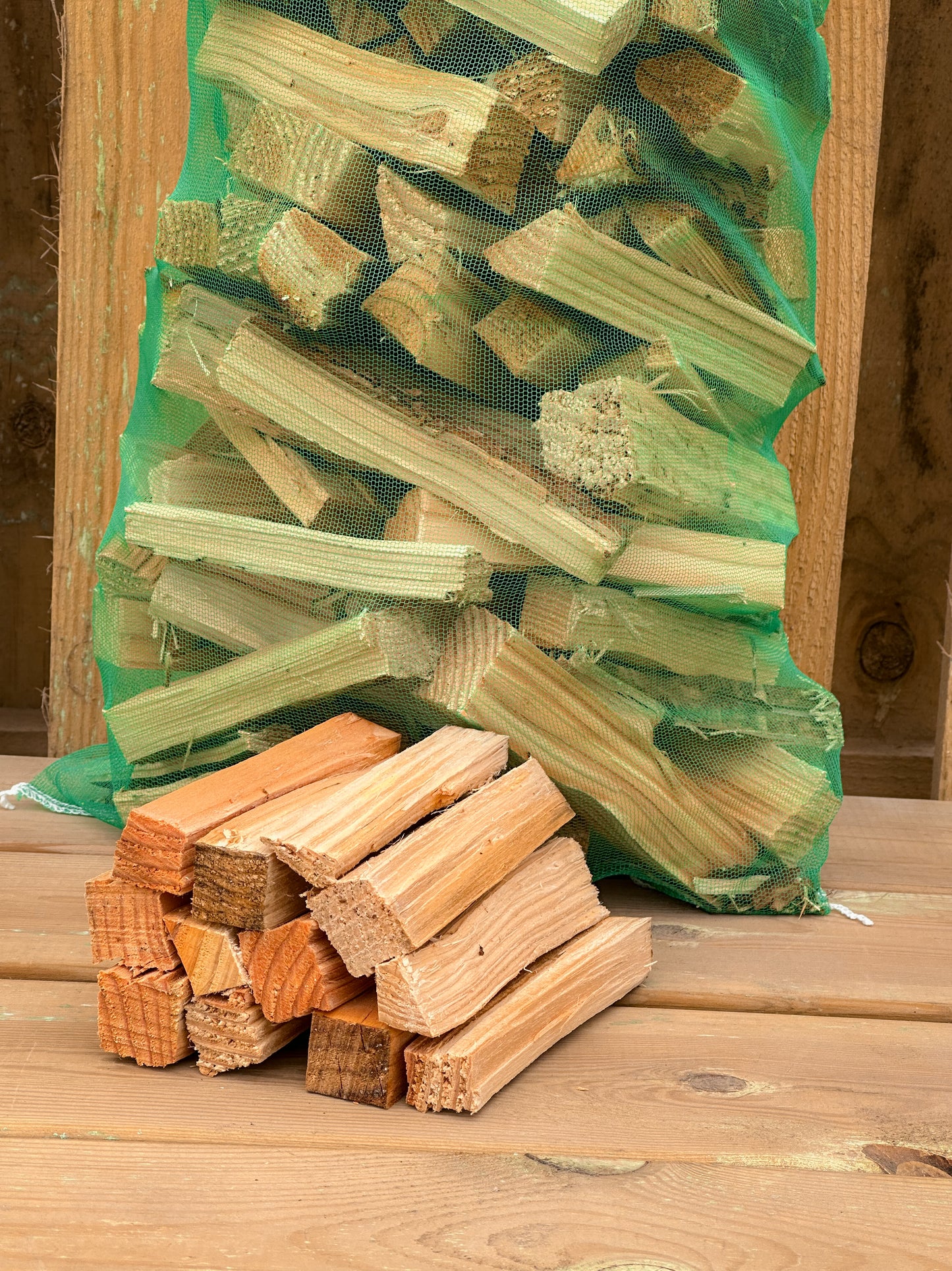 Here is a photo of our dry bags of kindling white pairs perfectly with our seasoned and kiln dried firewood. Small stack of kindling infront of green bag against wooden background.